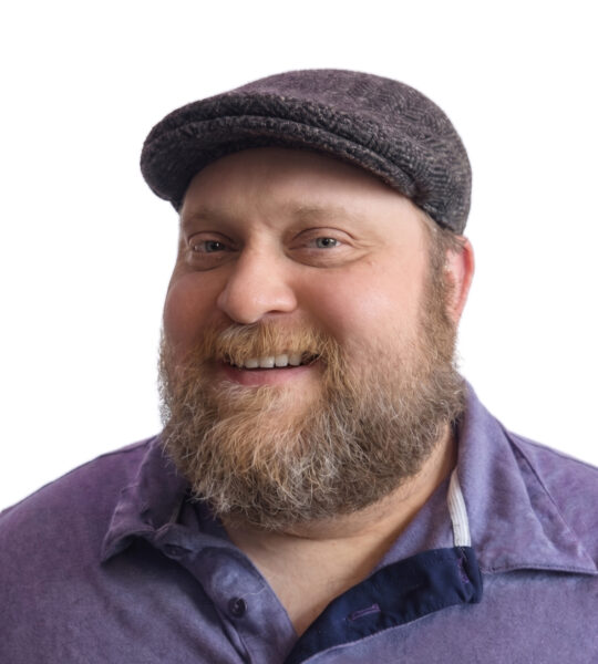 Professional headshot of a smiling man with a beard wearing a dark-colored shirt against a blurred background.