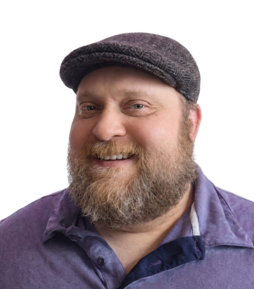 Professional headshot of a smiling man with a beard wearing a dark-colored shirt against a blurred background.