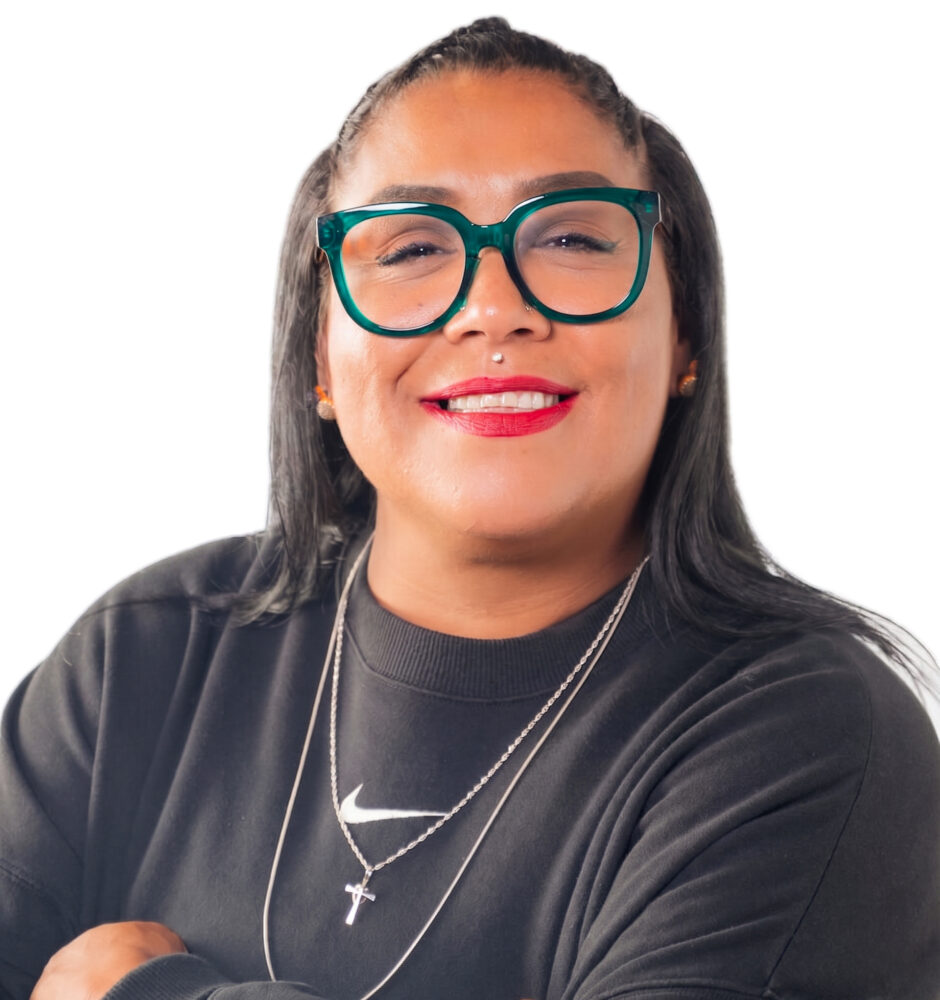 Professional headshot of a smiling woman wearing teal glasses and a black blazer against a white background.